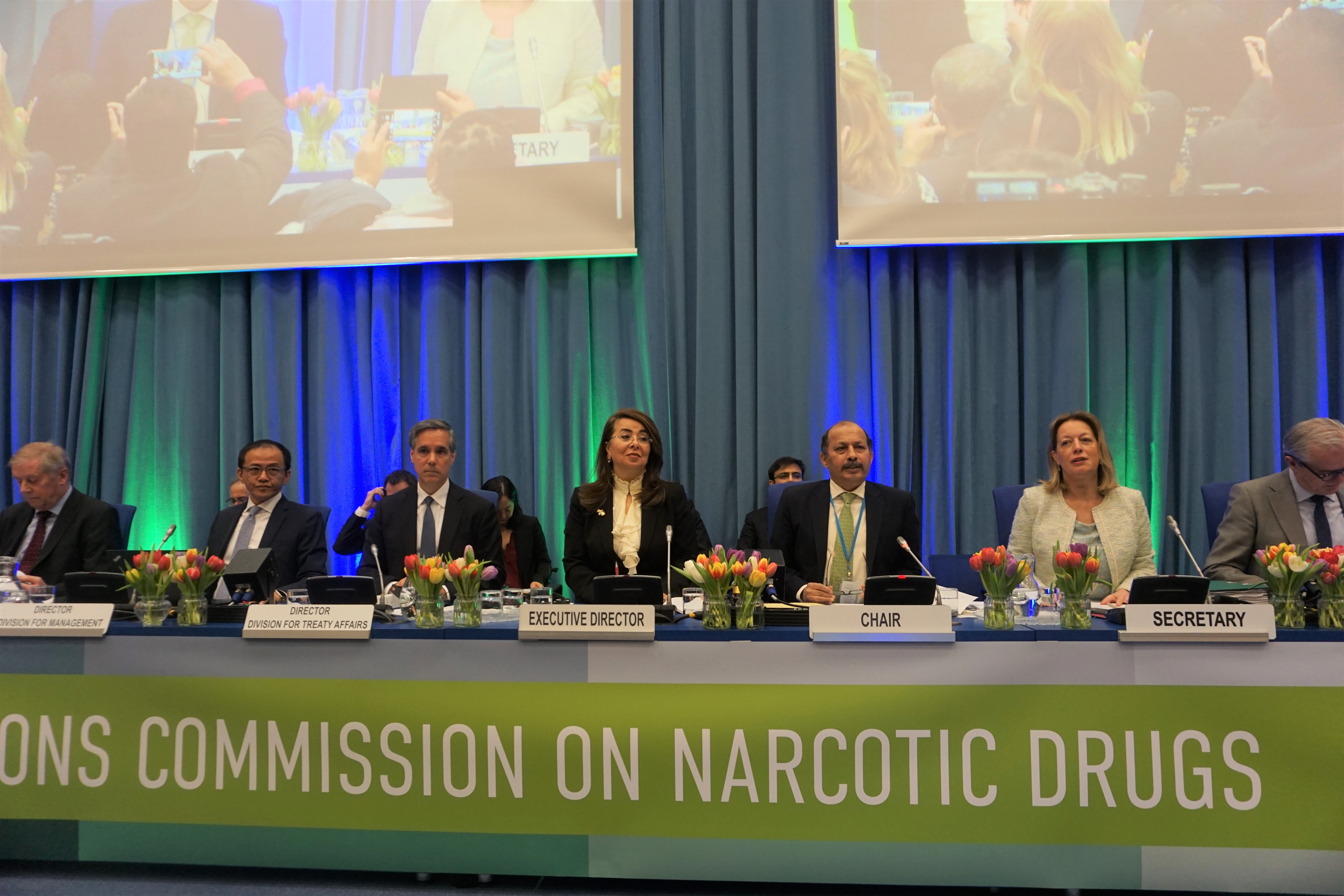 Speakers in formal attire sitting on the podium during the session of the Commission on Narcotic Drugs. The UNODC Executive Director was sitting in the middle of the picture with the Chair and the Secretary on the right side.