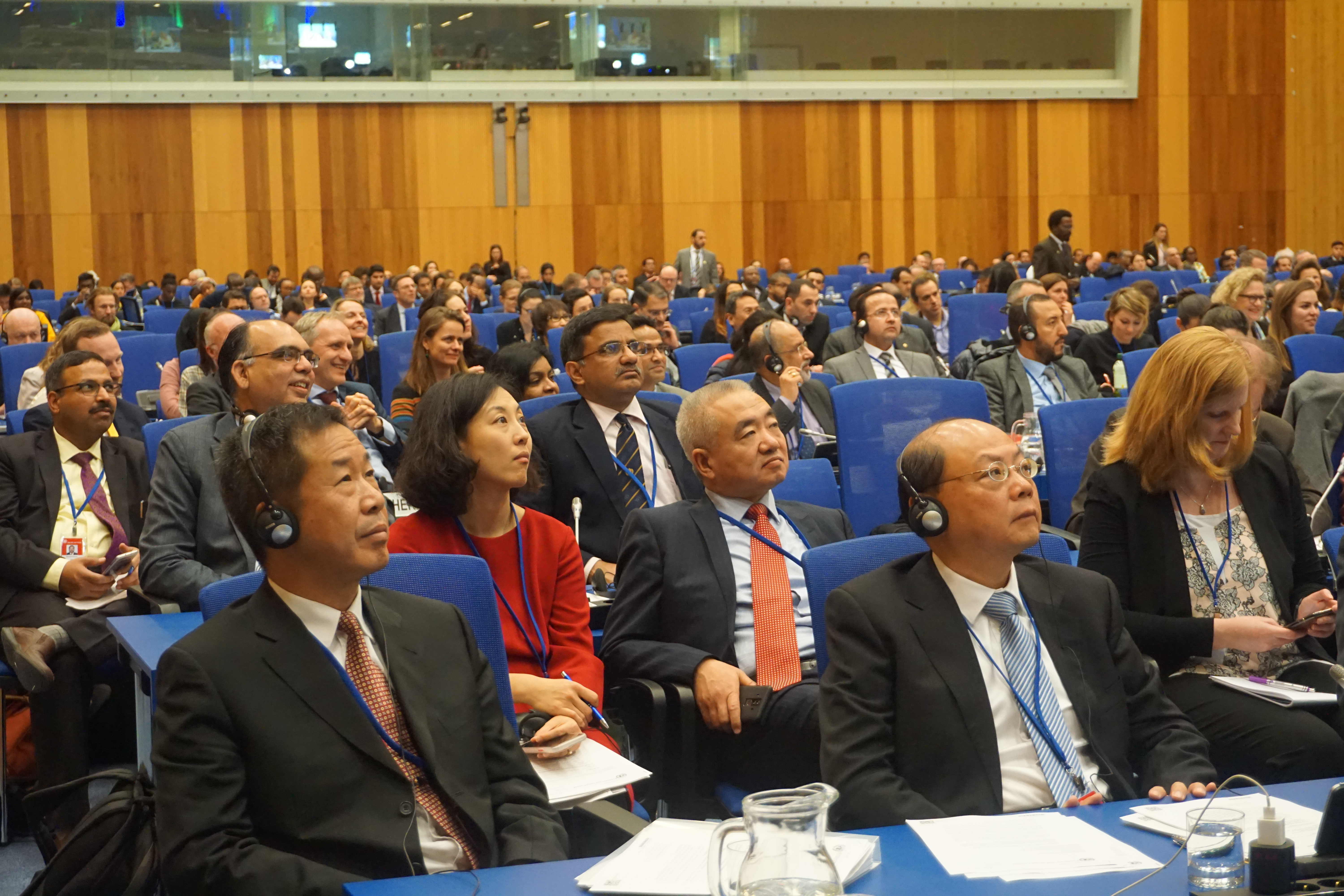 Participantes en vestimenta formal, sentados en una gran sala de conferencias, escuchando muy atentamente a los oradores.