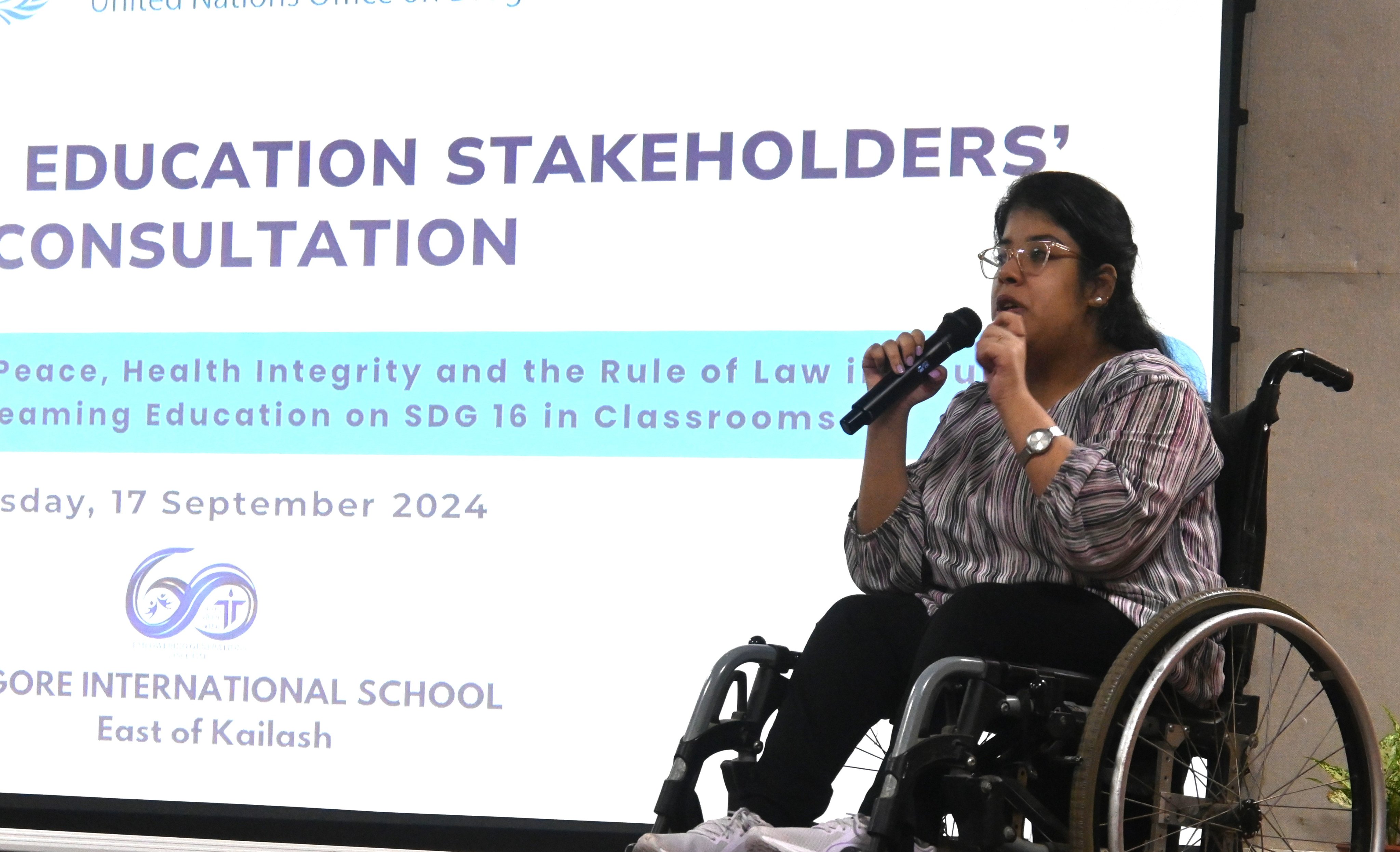 A girl with disabilities seated on a wheelchair, holding up a microphone and addressing a workshop on stage.